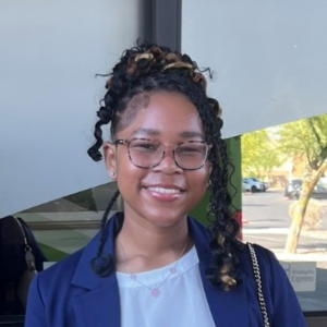 Elise Tyson in a navy blue blazer and white shirt smiling at the camera in front of an outdoor backdrop while wearing dark framed eye glasses