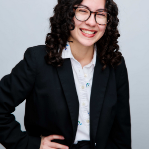 Isabella Whalen in a black blazer and white dress shirt smiling at the camera with her right hand on her hip while wearing dark framed eye glasses